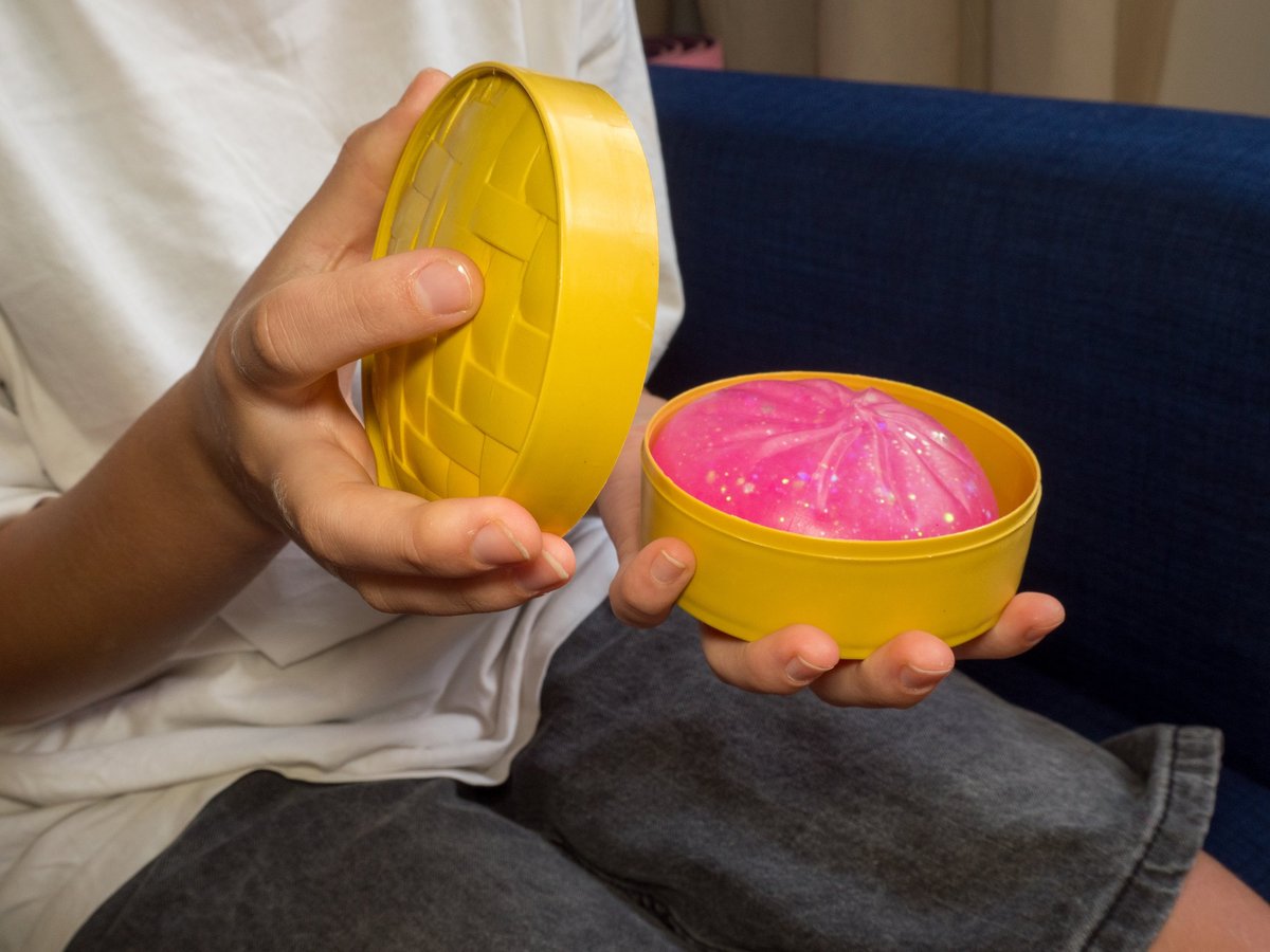 Close-up of a child's hands unboxing a pink glittery squishy dumpling toy from a yellow steamer basket